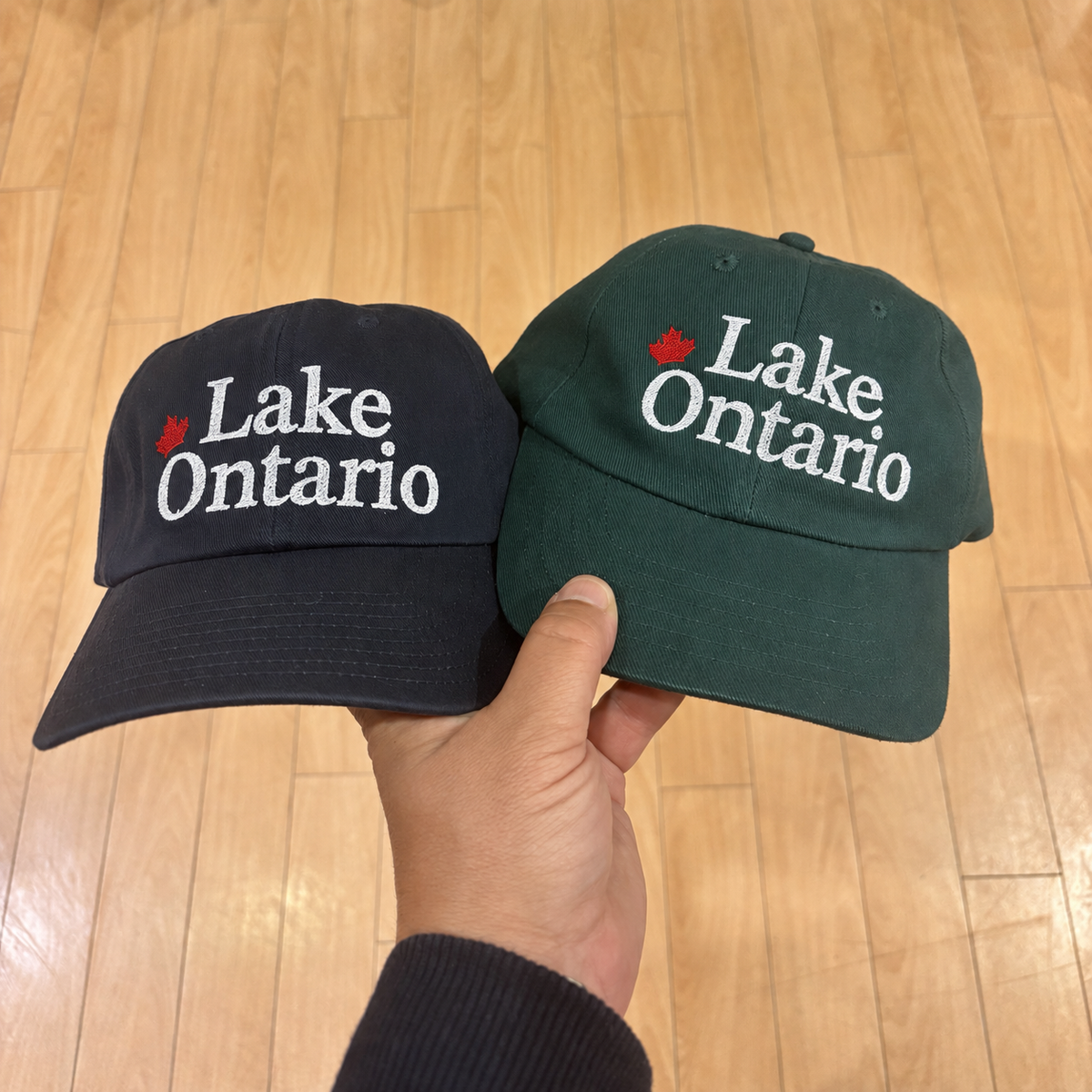 Navy and green Lake Ontario hats held side by side, showing white lettering and red maple leaves